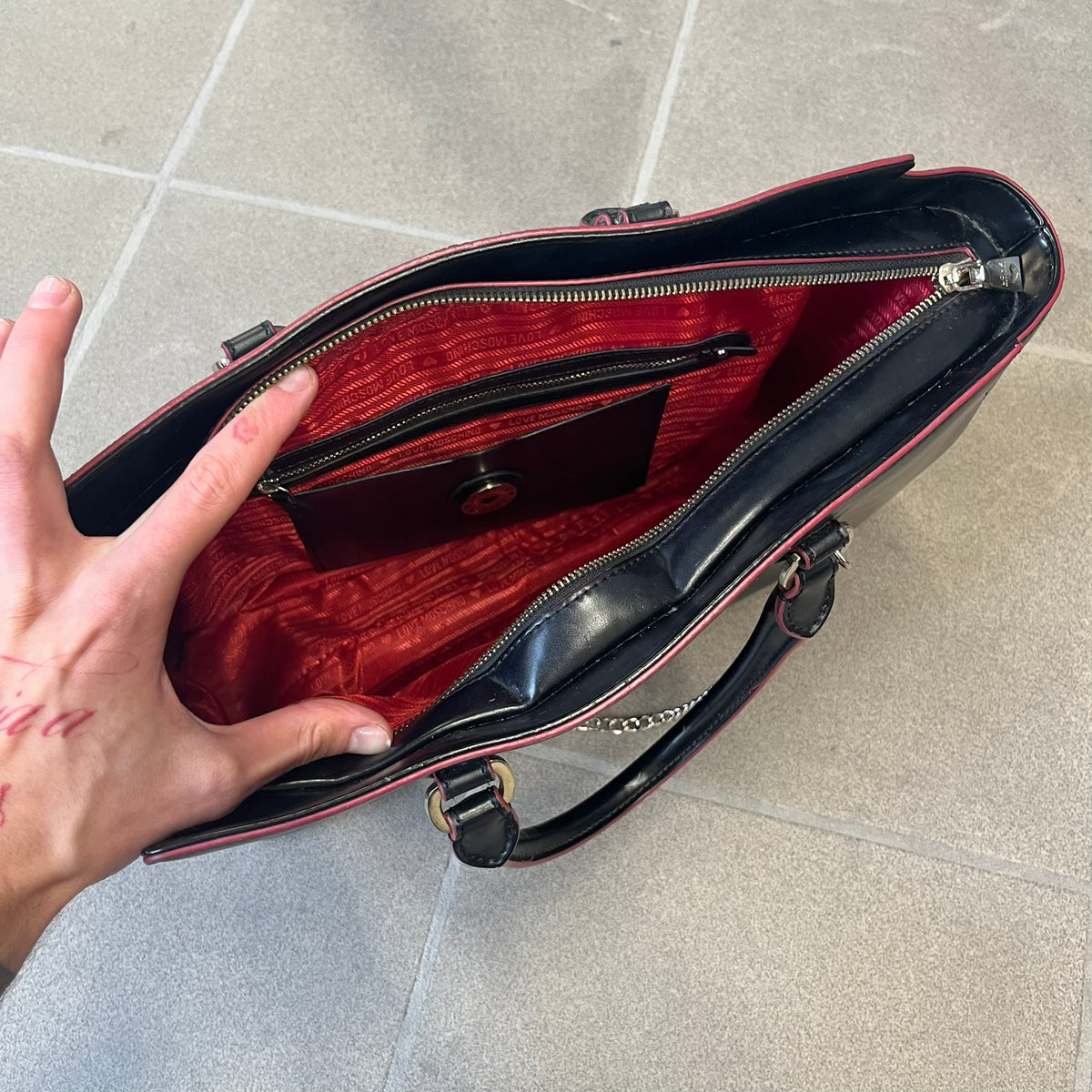 Love Moschino FW16 Heart Mirror Black Handbag is a sturdy tote-style handbag in black leather-look material with a red interior lining, two handles, a metal Love Moschino logo on the front, and branded buttons. It features a front pocket holding a heart-shaped mirror with a red back, attached to one handle by a chain.