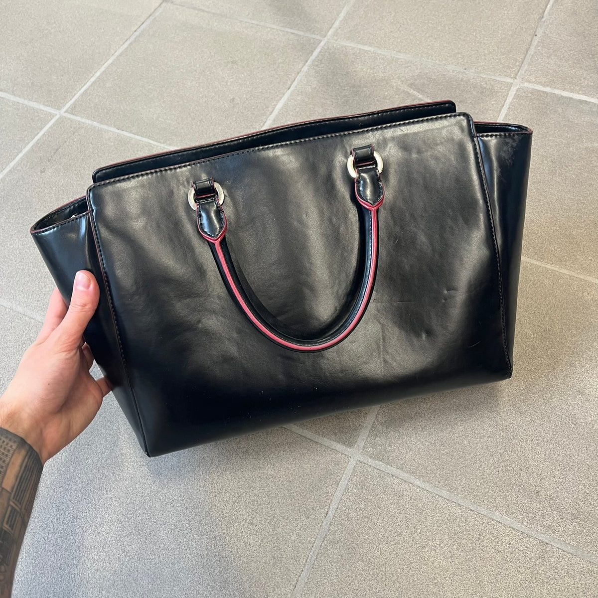 Love Moschino FW16 Heart Mirror Black Handbag is a sturdy tote-style handbag in black leather-look material with a red interior lining, two handles, a metal Love Moschino logo on the front, and branded buttons. It features a front pocket holding a heart-shaped mirror with a red back, attached to one handle by a chain.