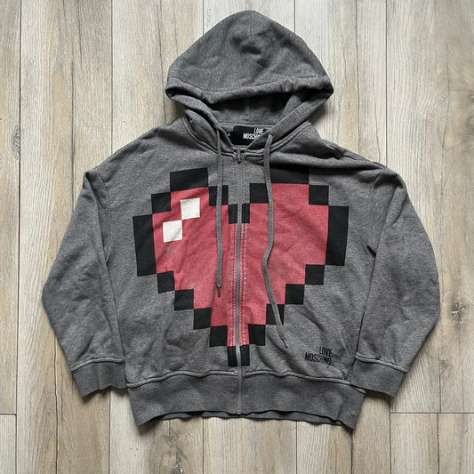 Love Moschino Pixel Heart Gray Hoodie is a gray zip-up hoodie featuring a large red pixel heart graphic on the front. It has slightly shorter-than-standard sleeves, long drawstrings, and a front zipper closure. The lower hem includes Love Moschino logo detailing, completing the branded design.