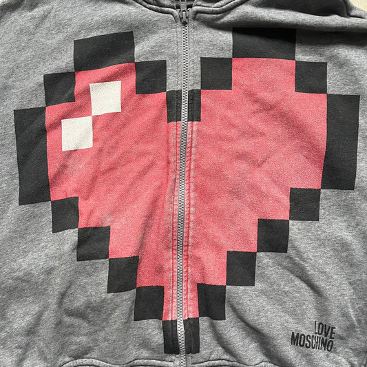 Love Moschino Pixel Heart Gray Hoodie is a gray zip-up hoodie featuring a large red pixel heart graphic on the front. It has slightly shorter-than-standard sleeves, long drawstrings, and a front zipper closure. The lower part includes Love Moschino logo detailing, completing the branded design.