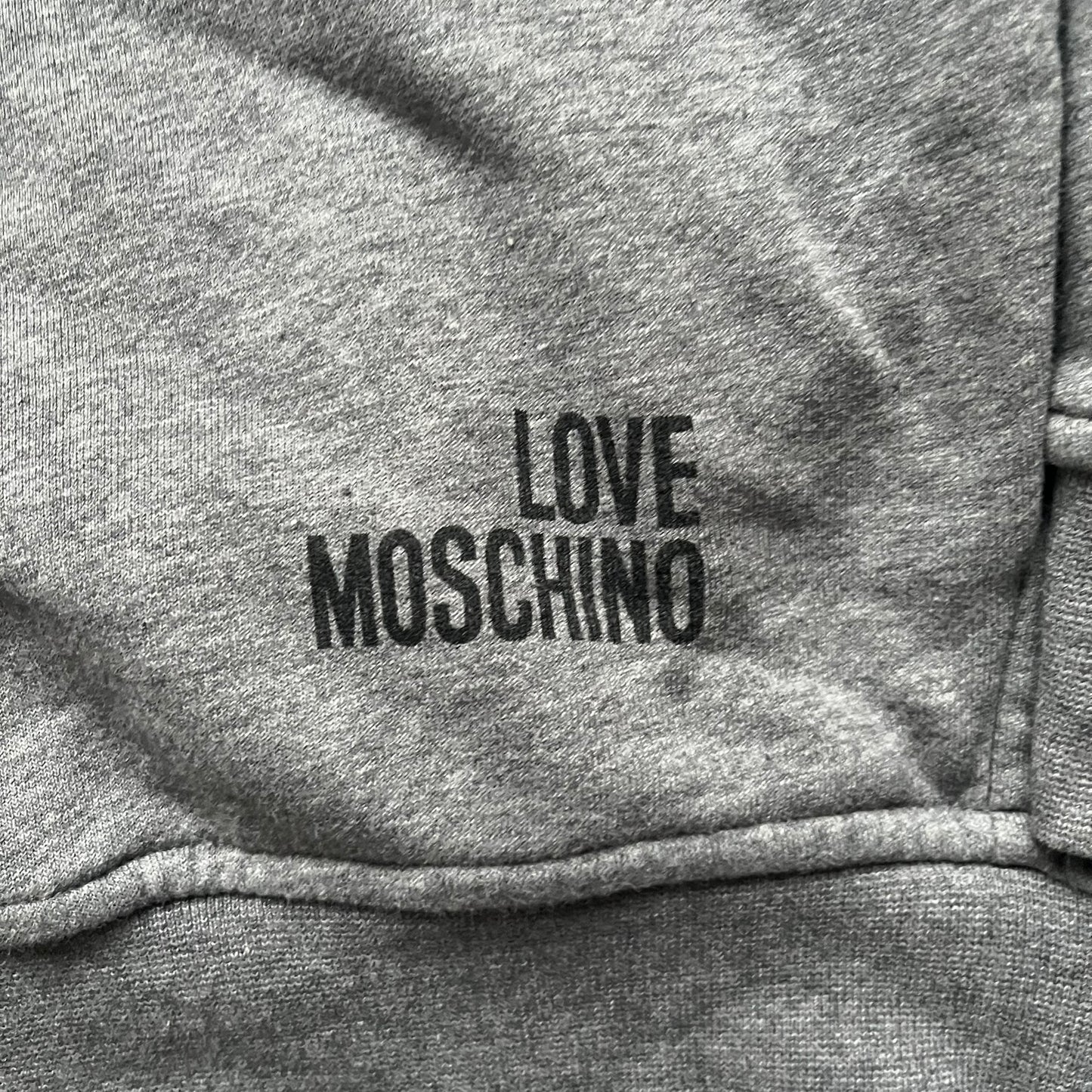 Love Moschino Pixel Heart Gray Hoodie is a gray zip-up hoodie featuring a large red pixel heart graphic on the front. It has slightly shorter-than-standard sleeves, long drawstrings, and a front zipper closure. The lower part includes Love Moschino logo detailing, completing the branded design.