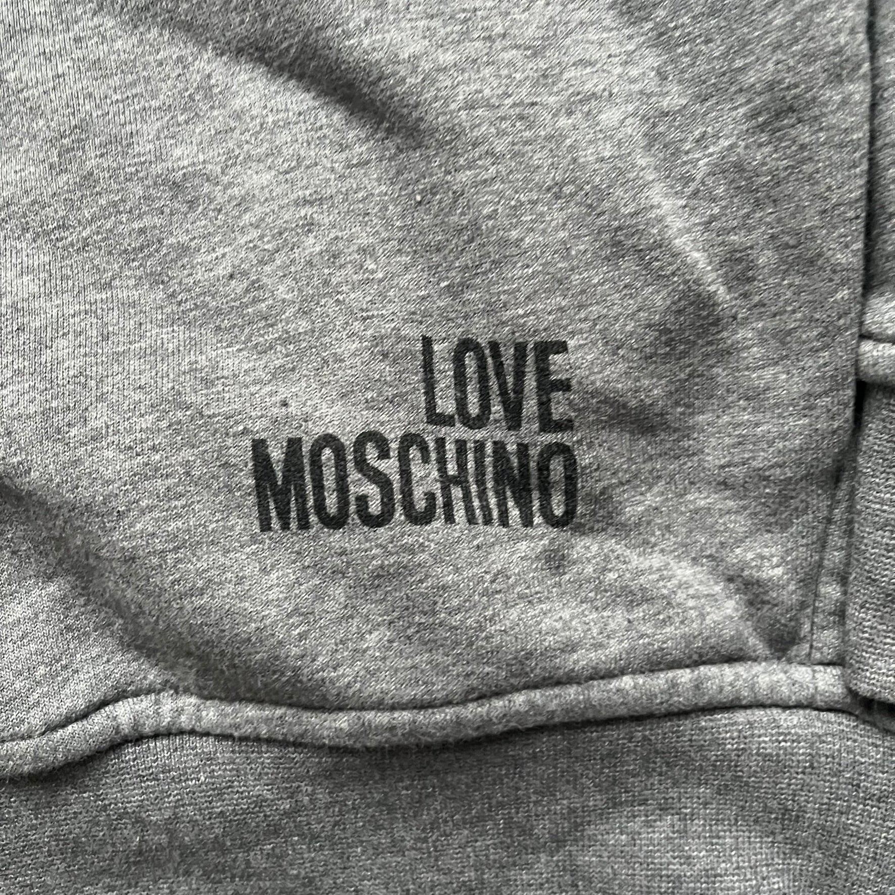 Love Moschino Pixel Heart Gray Hoodie is a gray zip-up hoodie featuring a large red pixel heart graphic on the front. It has slightly shorter-than-standard sleeves, long drawstrings, and a front zipper closure. The lower part includes Love Moschino logo detailing, completing the branded design.