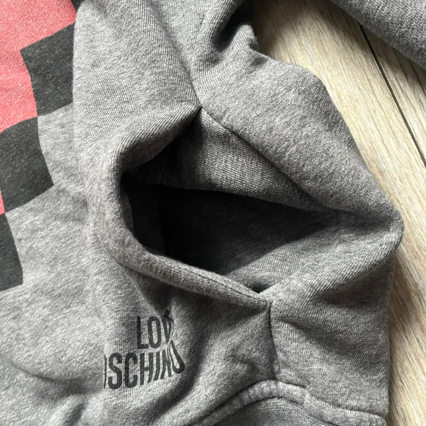 Love Moschino Pixel Heart Gray Hoodie is a gray zip-up hoodie featuring a large red pixel heart graphic on the front. It has slightly shorter-than-standard sleeves, long drawstrings, and a front zipper closure. The lower part includes Love Moschino logo detailing, completing the branded design.