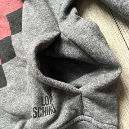 Love Moschino Pixel Heart Gray Hoodie is a gray zip-up hoodie featuring a large red pixel heart graphic on the front. It has slightly shorter-than-standard sleeves, long drawstrings, and a front zipper closure. The lower part includes Love Moschino logo detailing, completing the branded design.