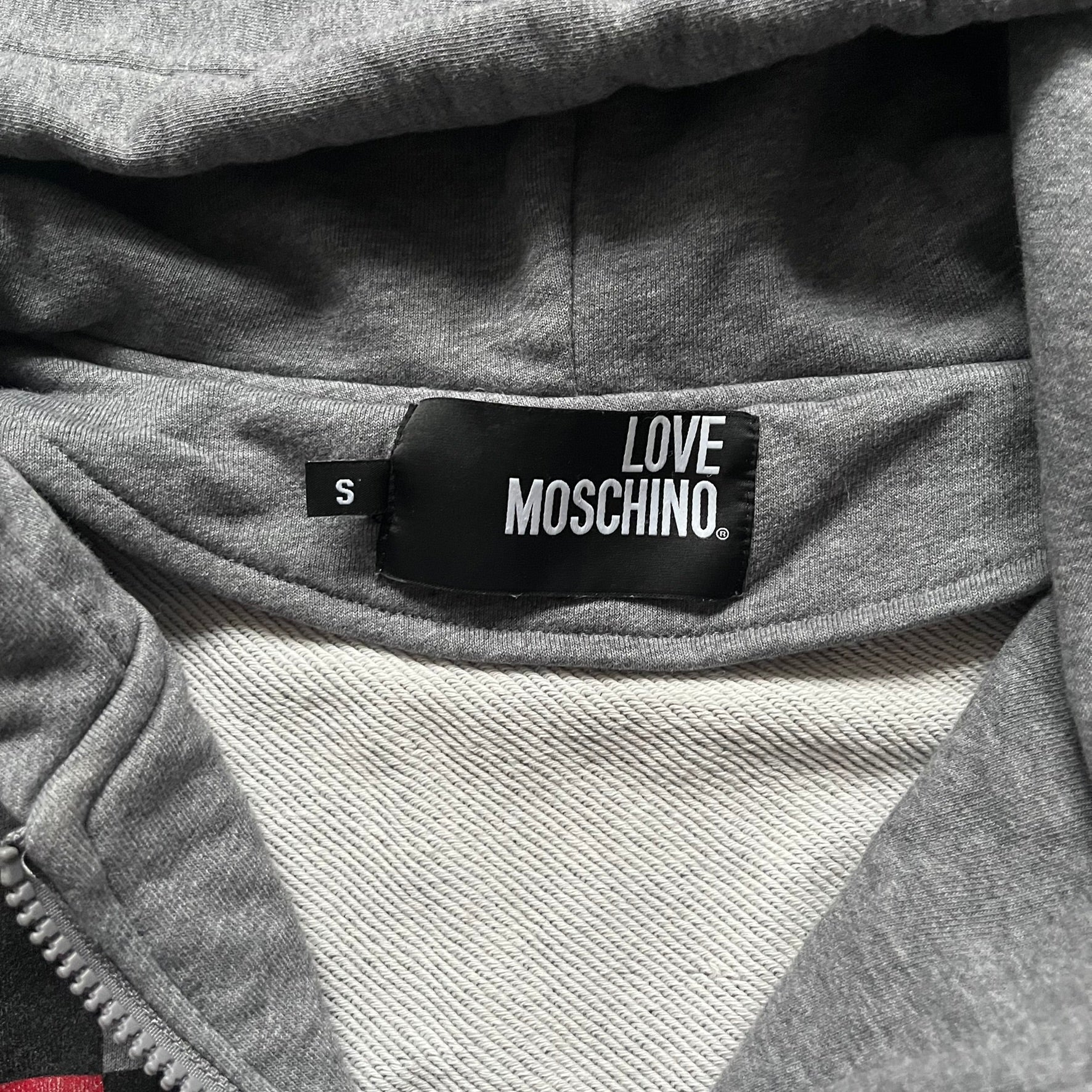Love Moschino Pixel Heart Gray Hoodie is a gray zip-up hoodie featuring a large red pixel heart graphic on the front. It has slightly shorter-than-standard sleeves, long drawstrings, and a front zipper closure. The lower part includes Love Moschino logo detailing, completing the branded design.