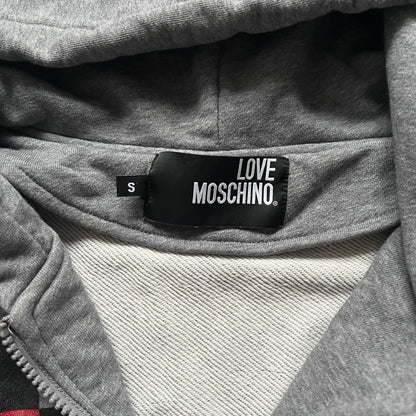 Love Moschino Pixel Heart Gray Hoodie is a gray zip-up hoodie featuring a large red pixel heart graphic on the front. It has slightly shorter-than-standard sleeves, long drawstrings, and a front zipper closure. The lower part includes Love Moschino logo detailing, completing the branded design.