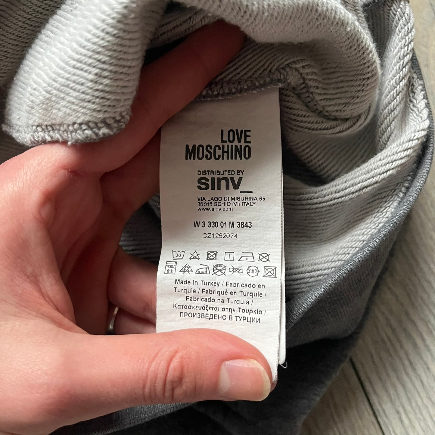 Love Moschino Pixel Heart Gray Hoodie is a gray zip-up hoodie featuring a large red pixel heart graphic on the front. It has slightly shorter-than-standard sleeves, long drawstrings, and a front zipper closure. The lower part includes Love Moschino logo detailing, completing the branded design.