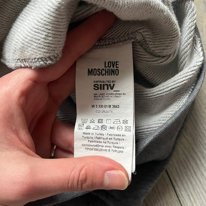 Love Moschino Pixel Heart Gray Hoodie is a gray zip-up hoodie featuring a large red pixel heart graphic on the front. It has slightly shorter-than-standard sleeves, long drawstrings, and a front zipper closure. The lower part includes Love Moschino logo detailing, completing the branded design.