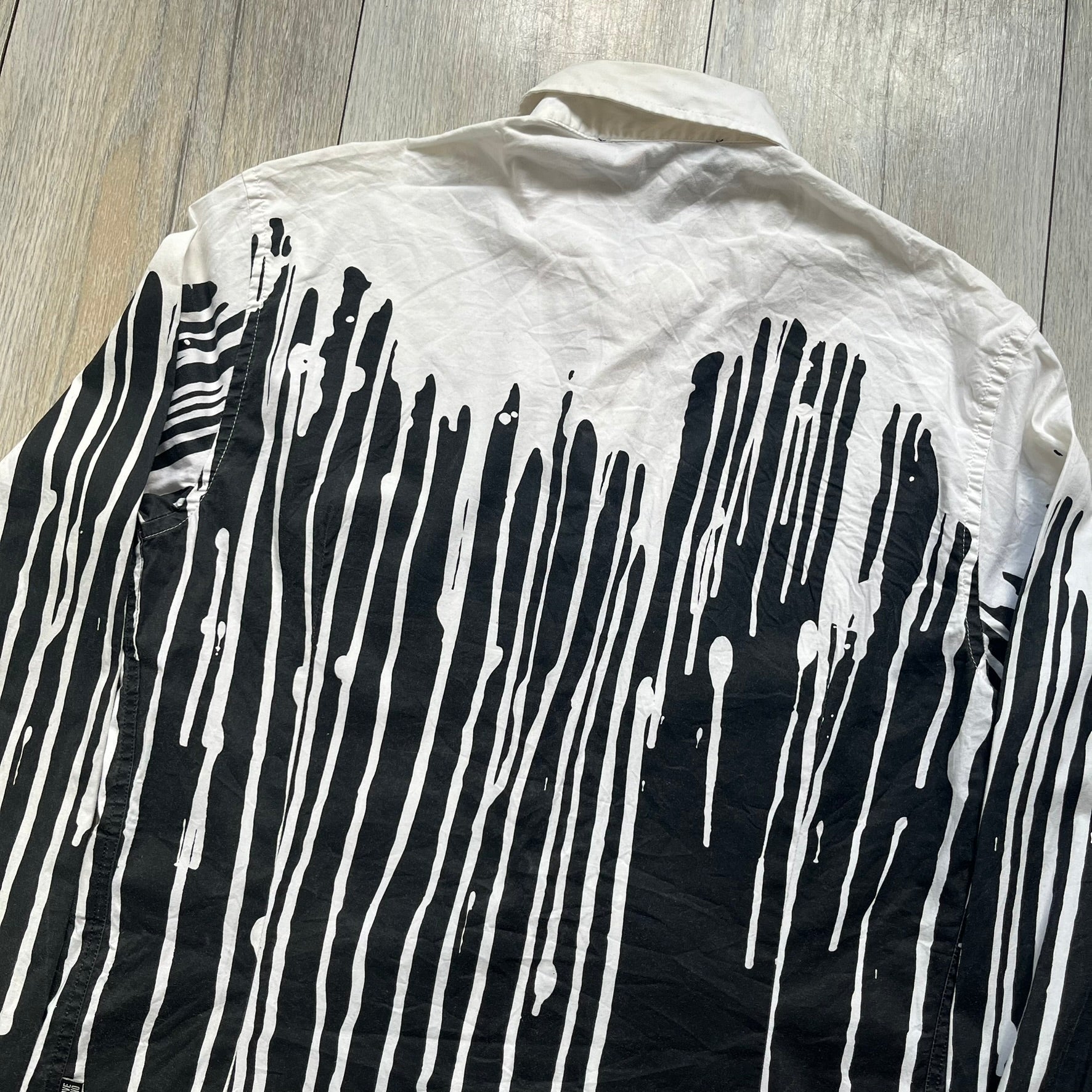 Love Moschino SS19 Dripping Paint Print Button-Up Shirt is a black and white shirt from the Spring/Summer 2019 collection. The upper portion is white, while the lower portion is black. The transition between the two sections features a dripping paint graphic extending downward from both sides into the black area. The button closure uses matching buttons, with white buttons on the white section and black buttons on the black section.
