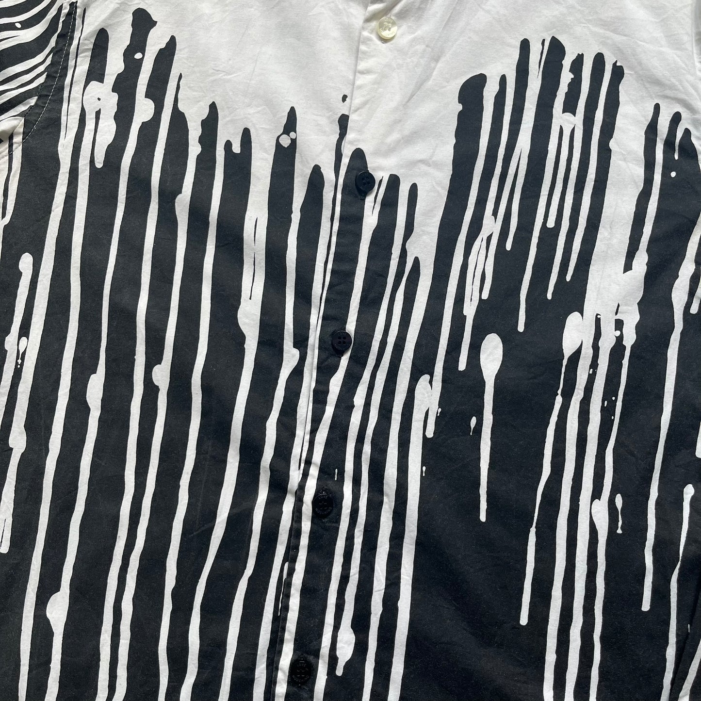 Love Moschino SS19 Dripping Paint Print Button-Up Shirt is a black and white shirt from the Spring/Summer 2019 collection. The upper portion is white, while the lower portion is black. The transition between the two sections features a dripping paint graphic extending downward from both sides into the black area. The button closure uses matching buttons, with white buttons on the white section and black buttons on the black section.

