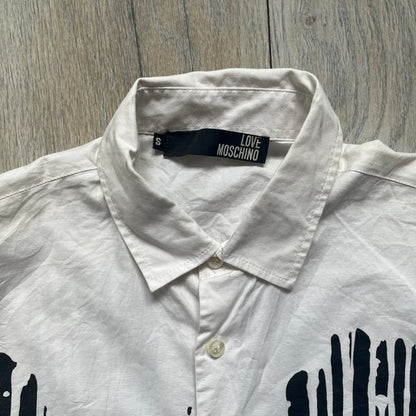 Love Moschino SS19 Dripping Paint Print Button-Up Shirt is a black and white shirt from the Spring/Summer 2019 collection. The upper portion is white, while the lower portion is black. The transition between the two sections features a dripping paint graphic extending downward from both sides into the black area. The button closure uses matching buttons, with white buttons on the white section and black buttons on the black section.
