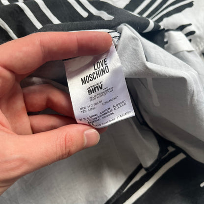 Love Moschino SS19 Dripping Paint Print Button-Up Shirt is a black and white shirt from the Spring/Summer 2019 collection. The upper portion is white, while the lower portion is black. The transition between the two sections features a dripping paint graphic extending downward from both sides into the black area. The button closure uses matching buttons, with white buttons on the white section and black buttons on the black section.
