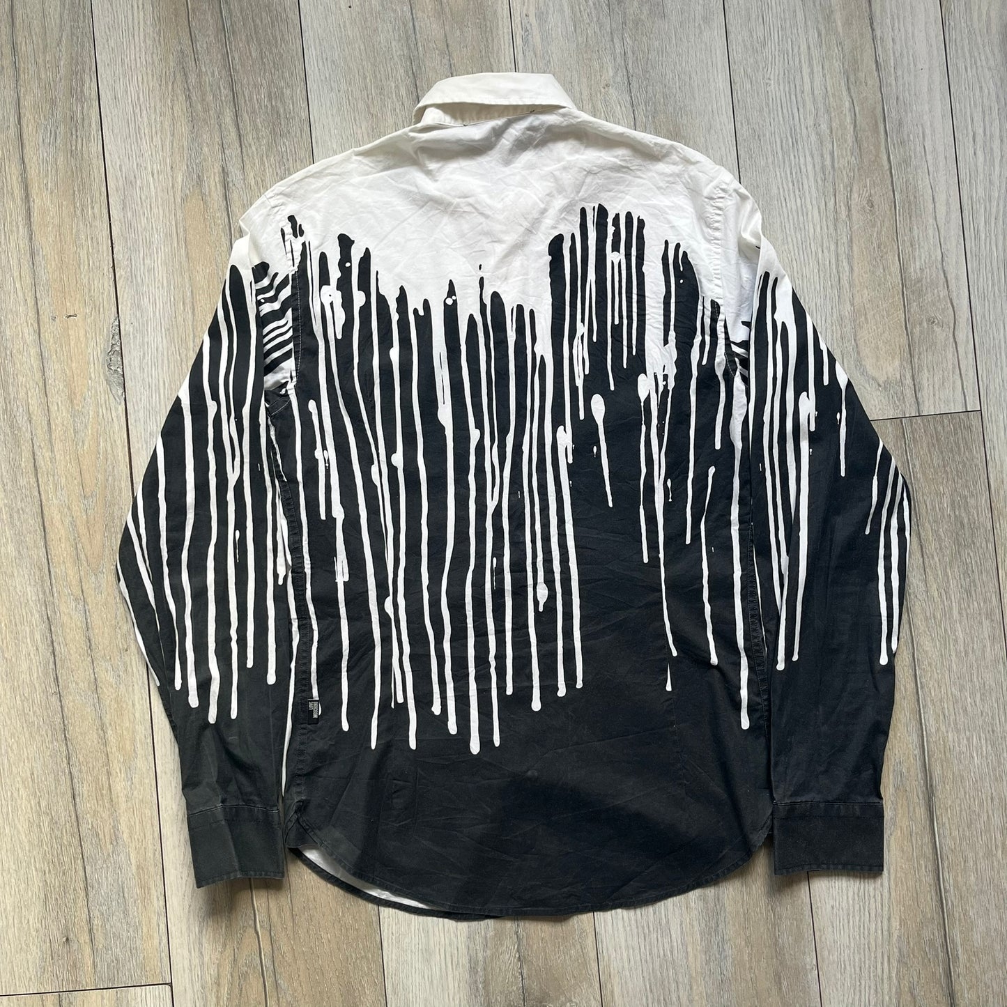 Love Moschino SS19 Dripping Paint Print Button-Up Shirt is a black and white shirt from the Spring/Summer 2019 collection. The upper portion is white, while the lower portion is black. The transition between the two sections features a dripping paint graphic extending downward from both sides into the black area. The button closure uses matching buttons, with white buttons on the white section and black buttons on the black section.
