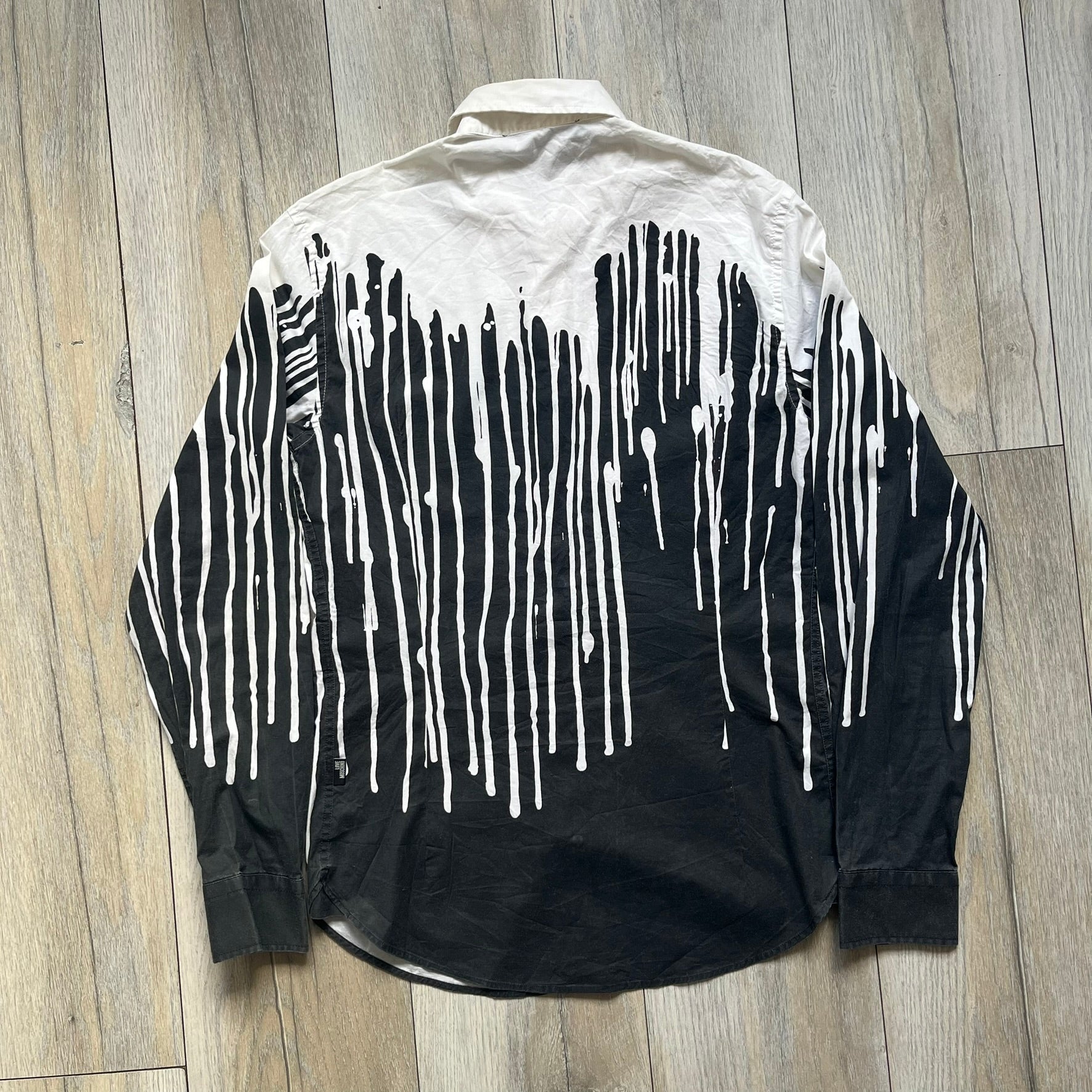 Love Moschino SS19 Dripping Paint Print Button-Up Shirt is a black and white shirt from the Spring/Summer 2019 collection. The upper portion is white, while the lower portion is black. The transition between the two sections features a dripping paint graphic extending downward from both sides into the black area. The button closure uses matching buttons, with white buttons on the white section and black buttons on the black section.
