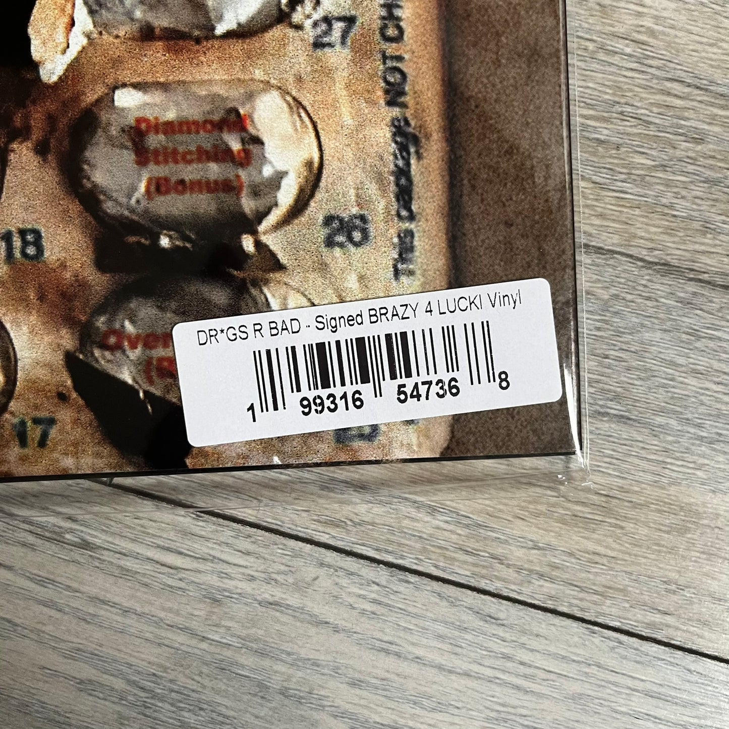 Lucki Drgs R Bad* 2026 Signed BRAZY 4 LUCKI Vinyl is a 2xLP release of Lucki’s fourth studio album, originally released on May 15, 2026. The set includes alternative front and back artwork, a widespine jacket, and printed inner sleeves with different graphics for each record. The vinyl records are transparent orange with black smoke effects throughout. The outer jacket features artwork of a girl shaving her head with the DRB logo cut into her hair. This copy is signed by Lucki in the upper left corner with 