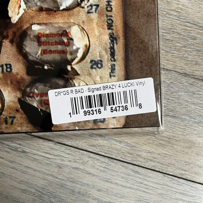 Lucki Drgs R Bad* 2026 Signed BRAZY 4 LUCKI Vinyl is a 2xLP release of Lucki’s fourth studio album, originally released on May 15, 2026. The set includes alternative front and back artwork, a widespine jacket, and printed inner sleeves with different graphics for each record. The vinyl records are transparent orange with black smoke effects throughout. The outer jacket features artwork of a girl shaving her head with the DRB logo cut into her hair. This copy is signed by Lucki in the upper left corner with 