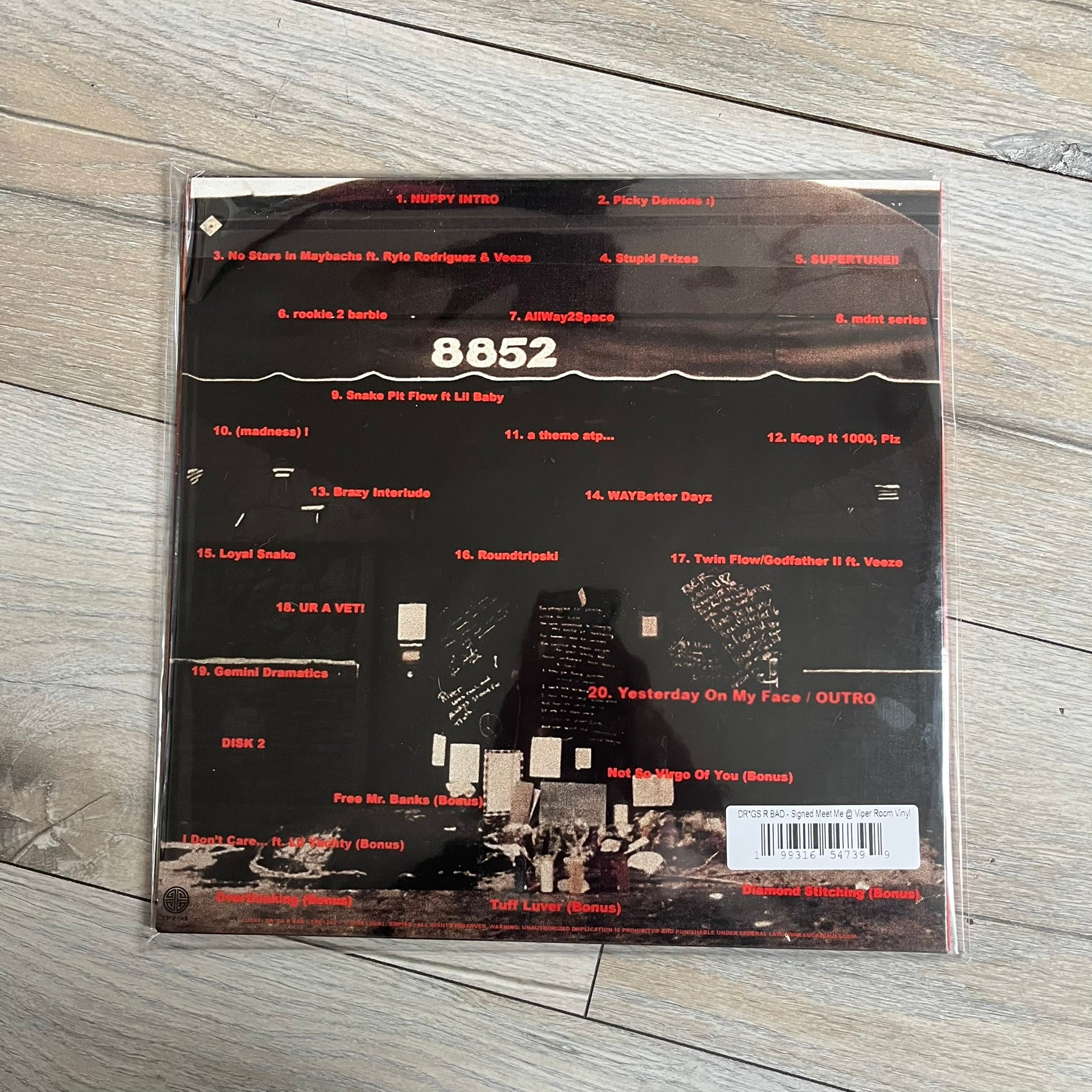 Lucki Dr*gs R Bad 2026 Signed Meet Me @ Viper Room Vinyl is a 2xLP release of Lucki’s fourth studio album, originally released on May 15, 2026. The set includes alternative front and back artwork, a widespine jacket, and printed inner sleeves with different graphics for each record. The vinyl records are transparent black with black smoke effects throughout. The outer jacket features artwork of a person drowning in a pool filled with money. This copy is signed by Lucki in the upper left corner with the insc