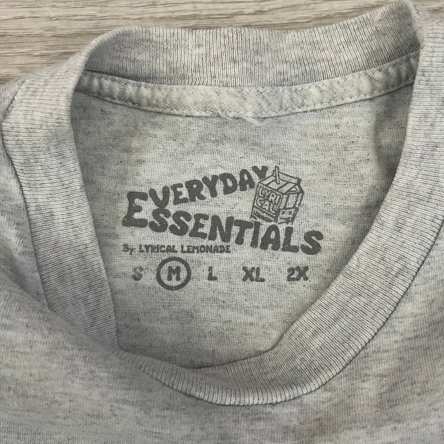 Lyrical Lemonade Everyday Essentials Gray T-Shirt in a gray colorway. Features a small Lyrical Lemonade carton graphic with an xP face printed on the left chest. The back displays a large Lyrical Lemonade logo with text and carton graphics in blue, yellow, and pink, from the Everyday Essentials line.