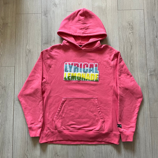 Lyrical Lemonade x Jordan hoodie in pink/coral, style code CW1122-670. Features a multicolor Lyrical Lemonade logo on the front in pink, yellow, blue, and green, designed with a textured, carpet-like finish and fluid graphic shape. Finished with an embroidered pink Jordan logo on the sleeve near the hem, a classic hood, and a front kangaroo pocket.