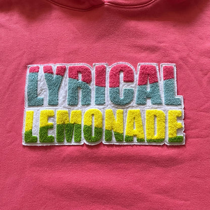Lyrical Lemonade x Jordan hoodie in pink/coral, style code CW1122-670. Features a multicolor Lyrical Lemonade logo on the front in pink, yellow, blue, and green, designed with a textured, carpet-like finish and fluid graphic shape. Finished with an embroidered pink Jordan logo on the sleeve near the hem, a classic hood, and a front kangaroo pocket.