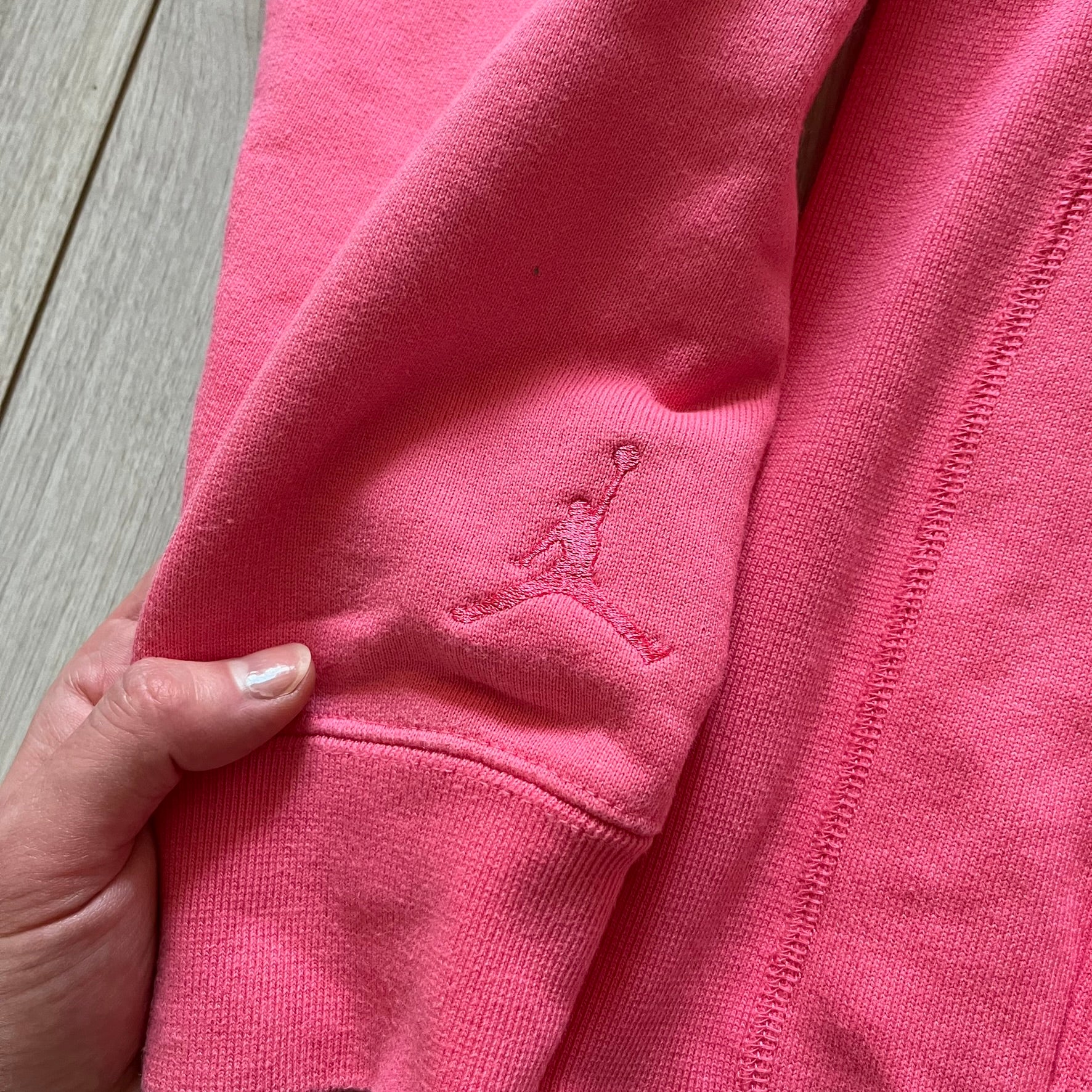 Lyrical Lemonade x Jordan hoodie in pink/coral, style code CW1122-670. Features a multicolor Lyrical Lemonade logo on the front in pink, yellow, blue, and green, designed with a textured, carpet-like finish and fluid graphic shape. Finished with an embroidered pink Jordan logo on the sleeve near the hem, a classic hood, and a front kangaroo pocket.