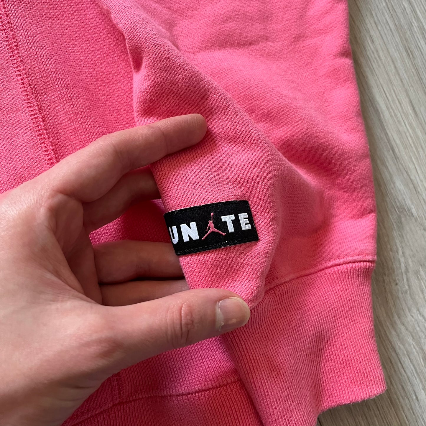 Lyrical Lemonade x Jordan hoodie in pink/coral, style code CW1122-670. Features a multicolor Lyrical Lemonade logo on the front in pink, yellow, blue, and green, designed with a textured, carpet-like finish and fluid graphic shape. Finished with an embroidered pink Jordan logo on the sleeve near the hem, a classic hood, and a front kangaroo pocket.