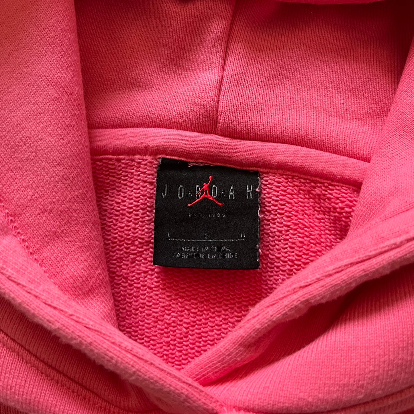 Lyrical Lemonade x Jordan hoodie in pink/coral, style code CW1122-670. Features a multicolor Lyrical Lemonade logo on the front in pink, yellow, blue, and green, designed with a textured, carpet-like finish and fluid graphic shape. Finished with an embroidered pink Jordan logo on the sleeve near the hem, a classic hood, and a front kangaroo pocket.