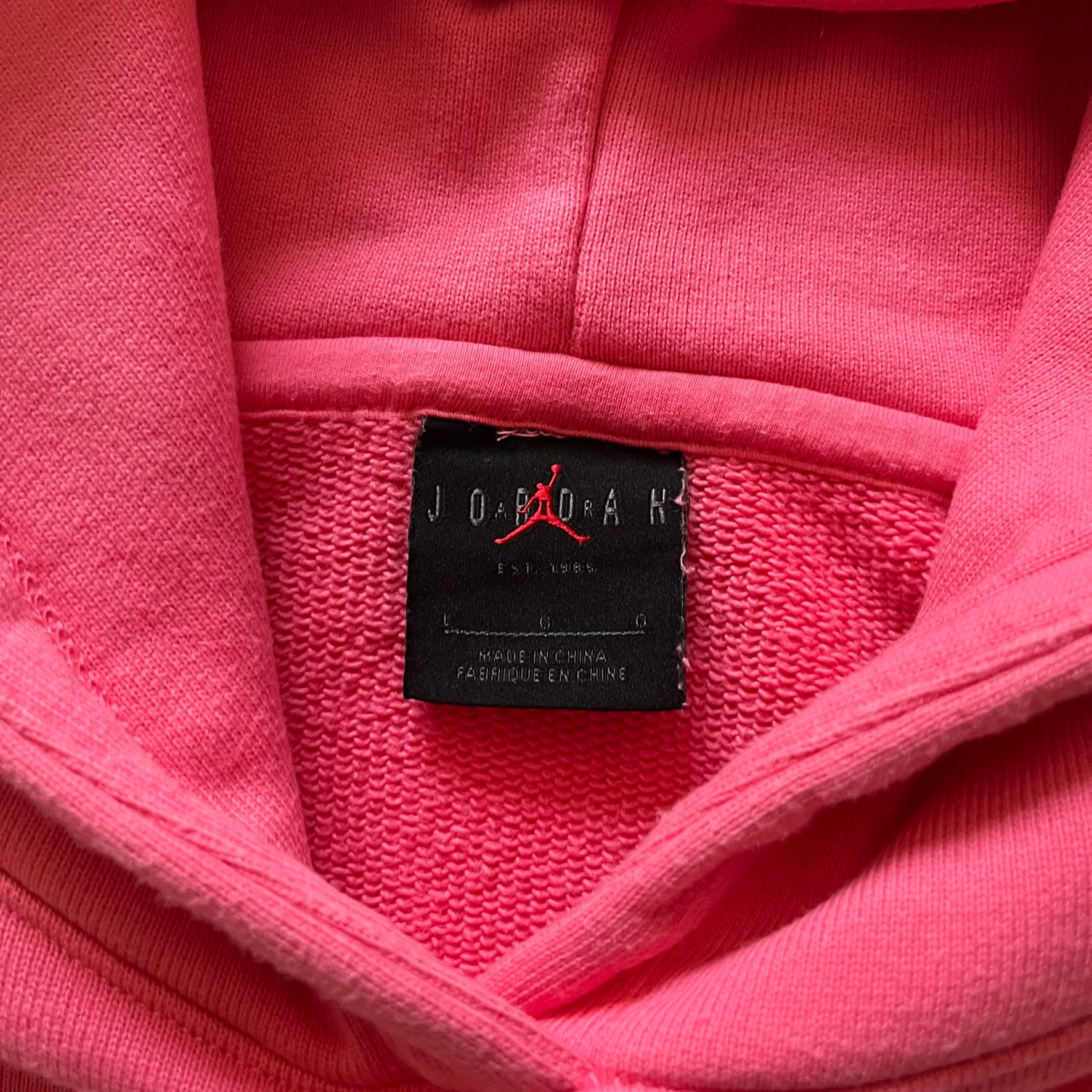 Lyrical Lemonade x Jordan hoodie in pink/coral, style code CW1122-670. Features a multicolor Lyrical Lemonade logo on the front in pink, yellow, blue, and green, designed with a textured, carpet-like finish and fluid graphic shape. Finished with an embroidered pink Jordan logo on the sleeve near the hem, a classic hood, and a front kangaroo pocket.
