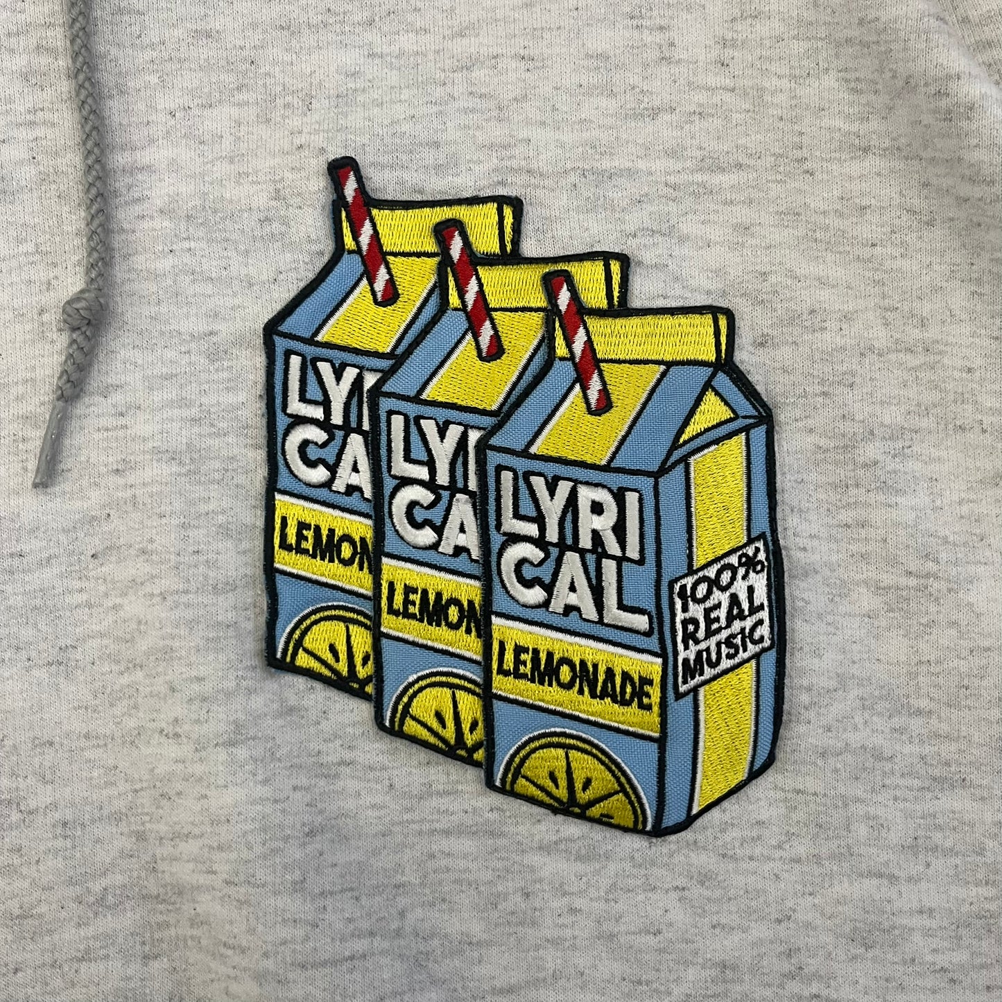 Champion x Lyrical Lemonade Grey Hoodie featuring a triple overlaping lemonade cartoon patch design on the front left chest. Made from a 50/50 cotton-polyester, this medium ash grey hoodie includes a two-ply hood, dyed-to-match drawcord, front pouch pocket, and embroidered Champion logo on the wrist.