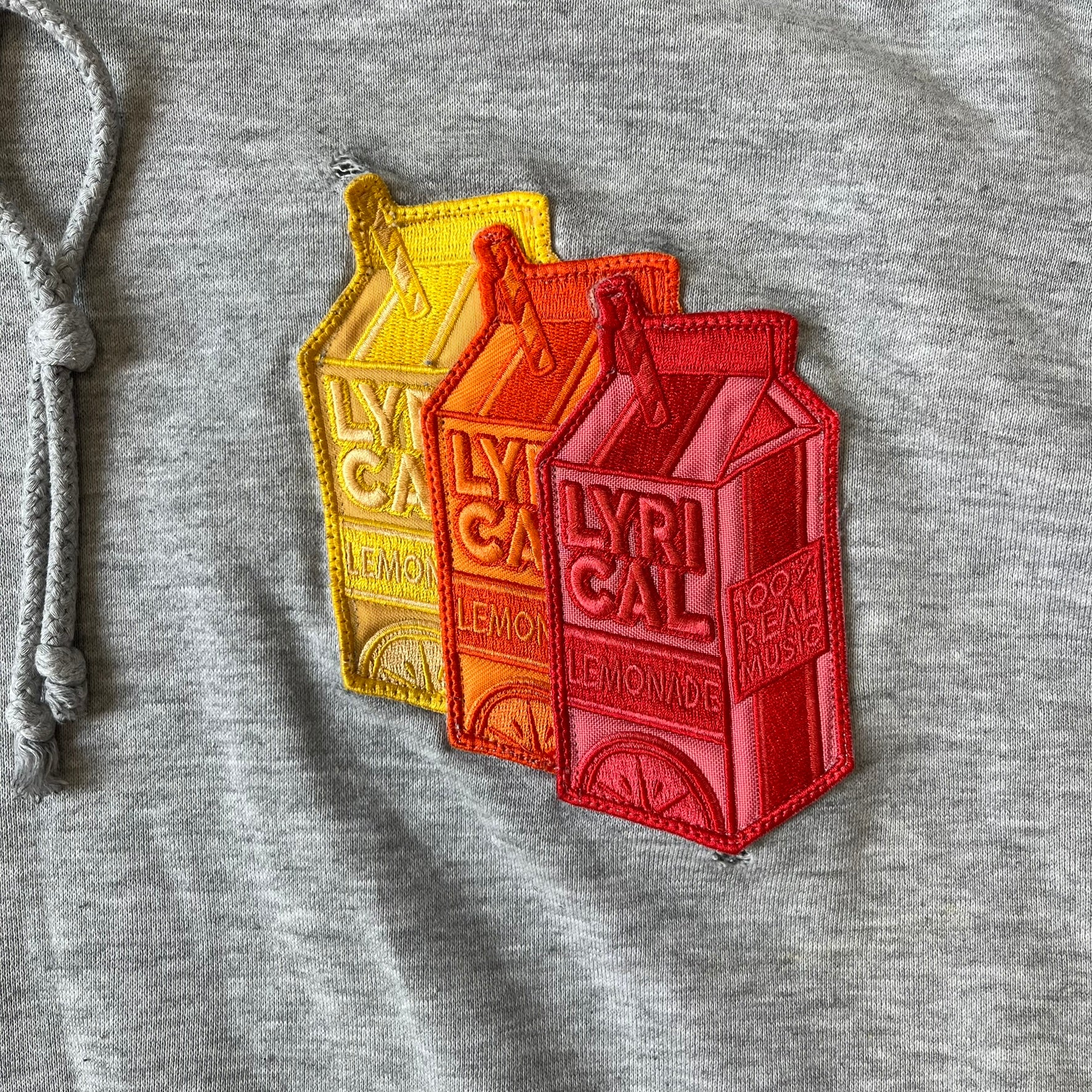 Lyrical Lemonade Triple Patch Gray Hoodie features a gray colorway with a layered triple lemonade box patch design on the left chest in red, yellow, and blue. Made from an 80/20 cotton-polyester blend, the hoodie includes a yellow Lyrical Lemonade neck tag.