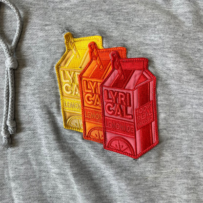 Lyrical Lemonade Triple Patch Gray Hoodie features a gray colorway with a layered triple lemonade box patch design on the left chest in red, yellow, and blue. Made from an 80/20 cotton-polyester blend, the hoodie includes a yellow Lyrical Lemonade neck tag.