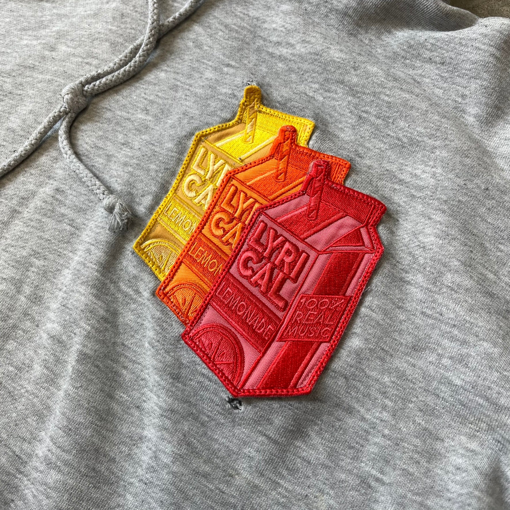 Lyrical Lemonade Triple Patch Gray Hoodie features a gray colorway with a layered triple lemonade box patch design on the left chest in red, yellow, and blue. Made from an 80/20 cotton-polyester blend, the hoodie includes a yellow Lyrical Lemonade neck tag.