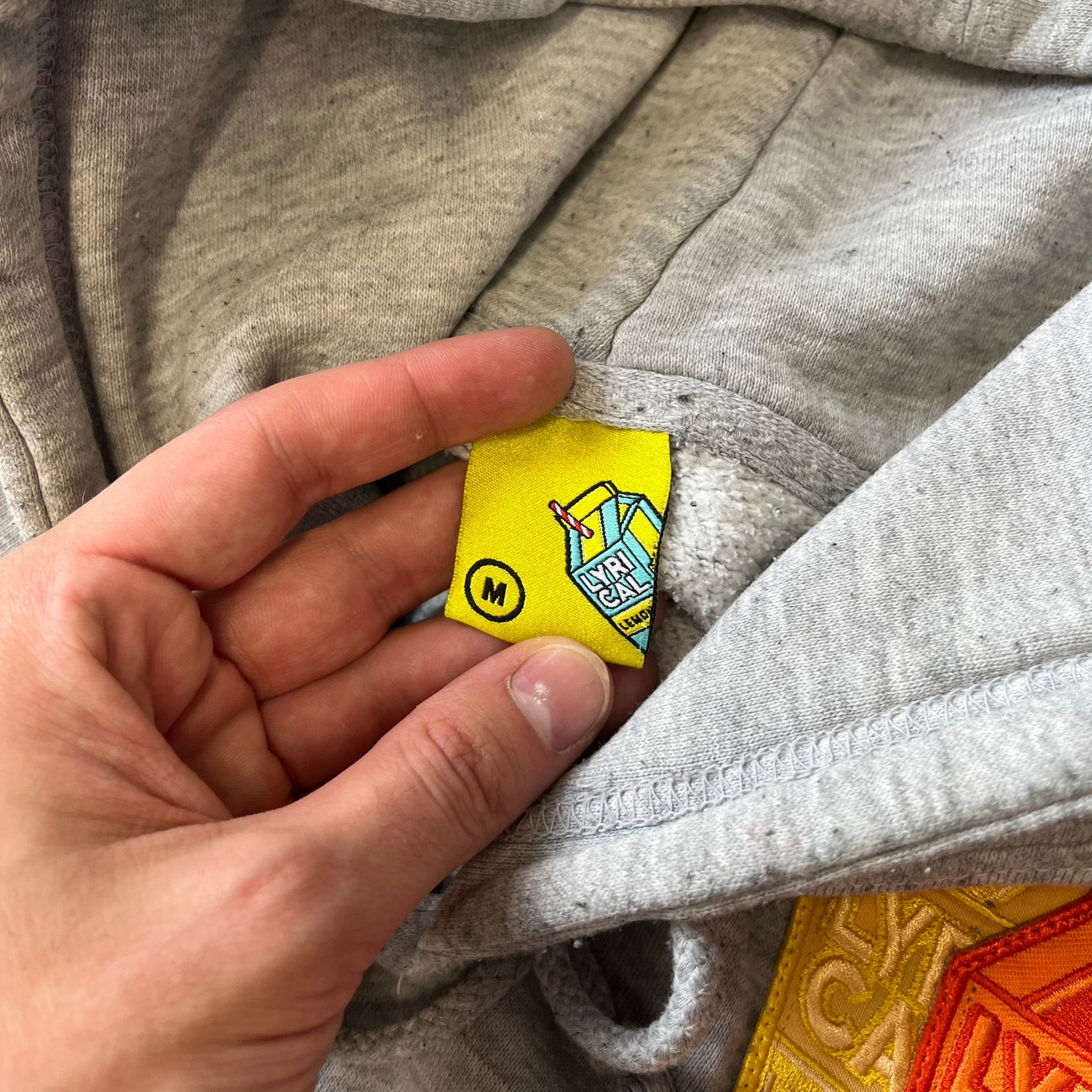 Lyrical Lemonade Triple Patch Gray Hoodie features a gray colorway with a layered triple lemonade box patch design on the left chest in red, yellow, and blue. Made from an 80/20 cotton-polyester blend, the hoodie includes a yellow Lyrical Lemonade neck tag.