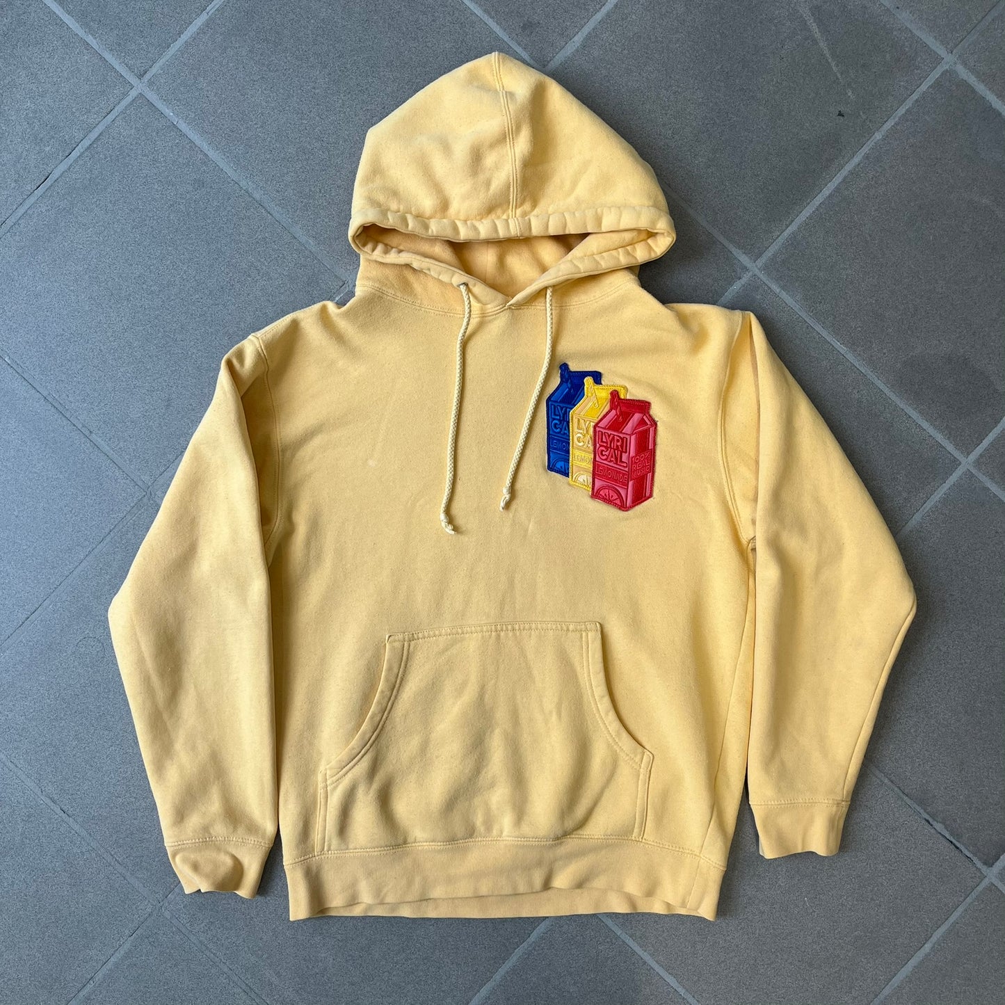 Lyrical Lemonade Triple Patch Yellow Hoodie features a yellow colorway with a layered triple lemonade box patch design on the left chest in red, yellow, and blue. Made from an 80/20 cotton-polyester blend, the hoodie includes a yellow Lyrical Lemonade neck tag.
