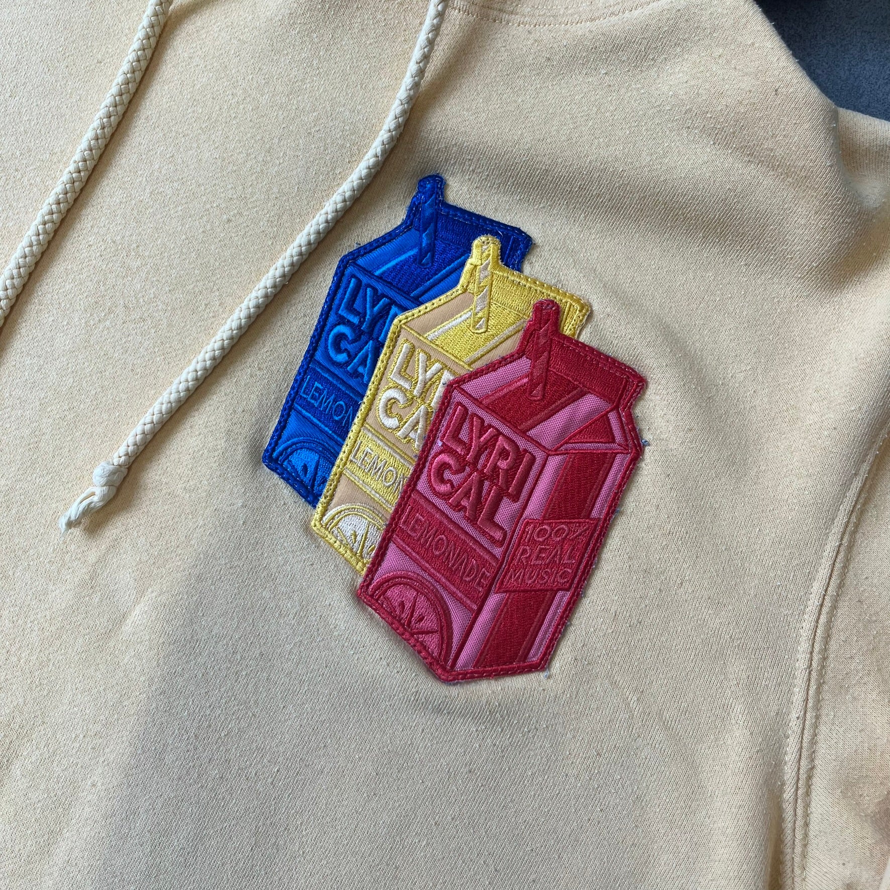 Lyrical Lemonade Triple Patch Yellow Hoodie features a yellow colorway with a layered triple lemonade box patch design on the left chest in red, yellow, and blue. Made from an 80/20 cotton-polyester blend, the hoodie includes a yellow Lyrical Lemonade neck tag.