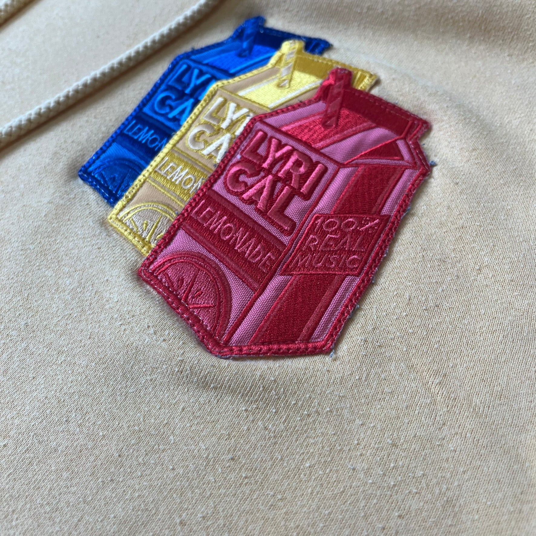 Lyrical Lemonade Triple Patch Yellow Hoodie features a yellow colorway with a layered triple lemonade box patch design on the left chest in red, yellow, and blue. Made from an 80/20 cotton-polyester blend, the hoodie includes a yellow Lyrical Lemonade neck tag.