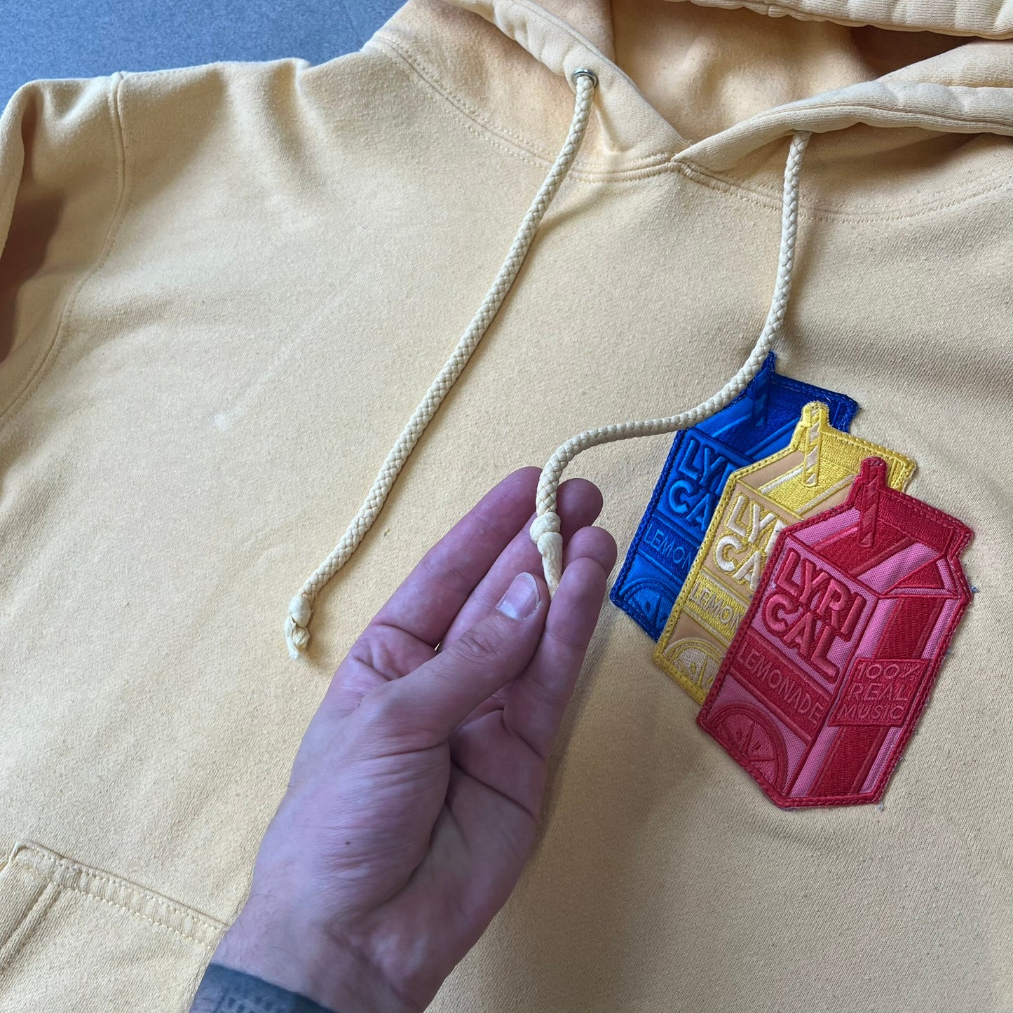 Lyrical Lemonade Triple Patch Yellow Hoodie features a yellow colorway with a layered triple lemonade box patch design on the left chest in red, yellow, and blue. Made from an 80/20 cotton-polyester blend, the hoodie includes a yellow Lyrical Lemonade neck tag.