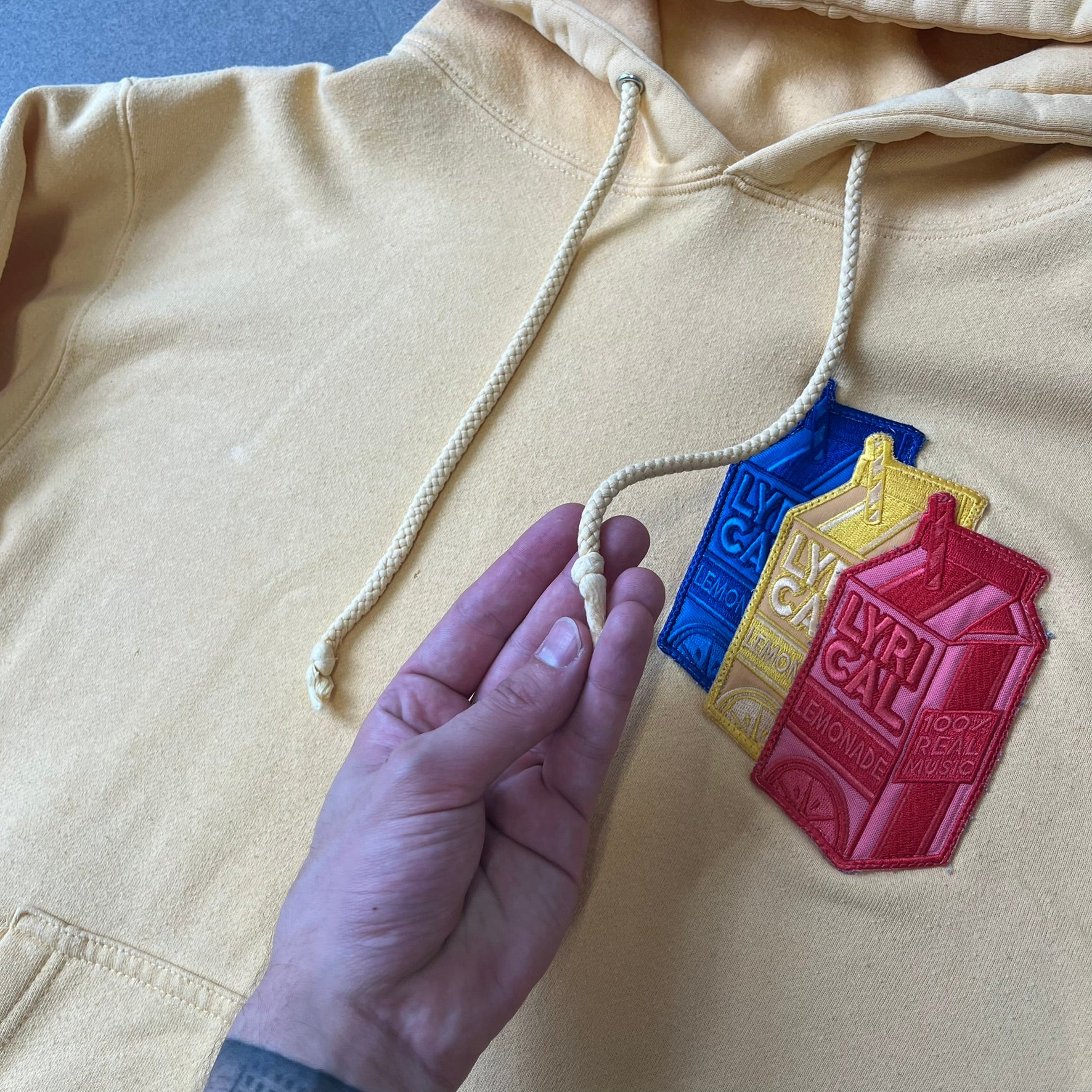Lyrical Lemonade Triple Patch Yellow Hoodie features a yellow colorway with a layered triple lemonade box patch design on the left chest in red, yellow, and blue. Made from an 80/20 cotton-polyester blend, the hoodie includes a yellow Lyrical Lemonade neck tag.