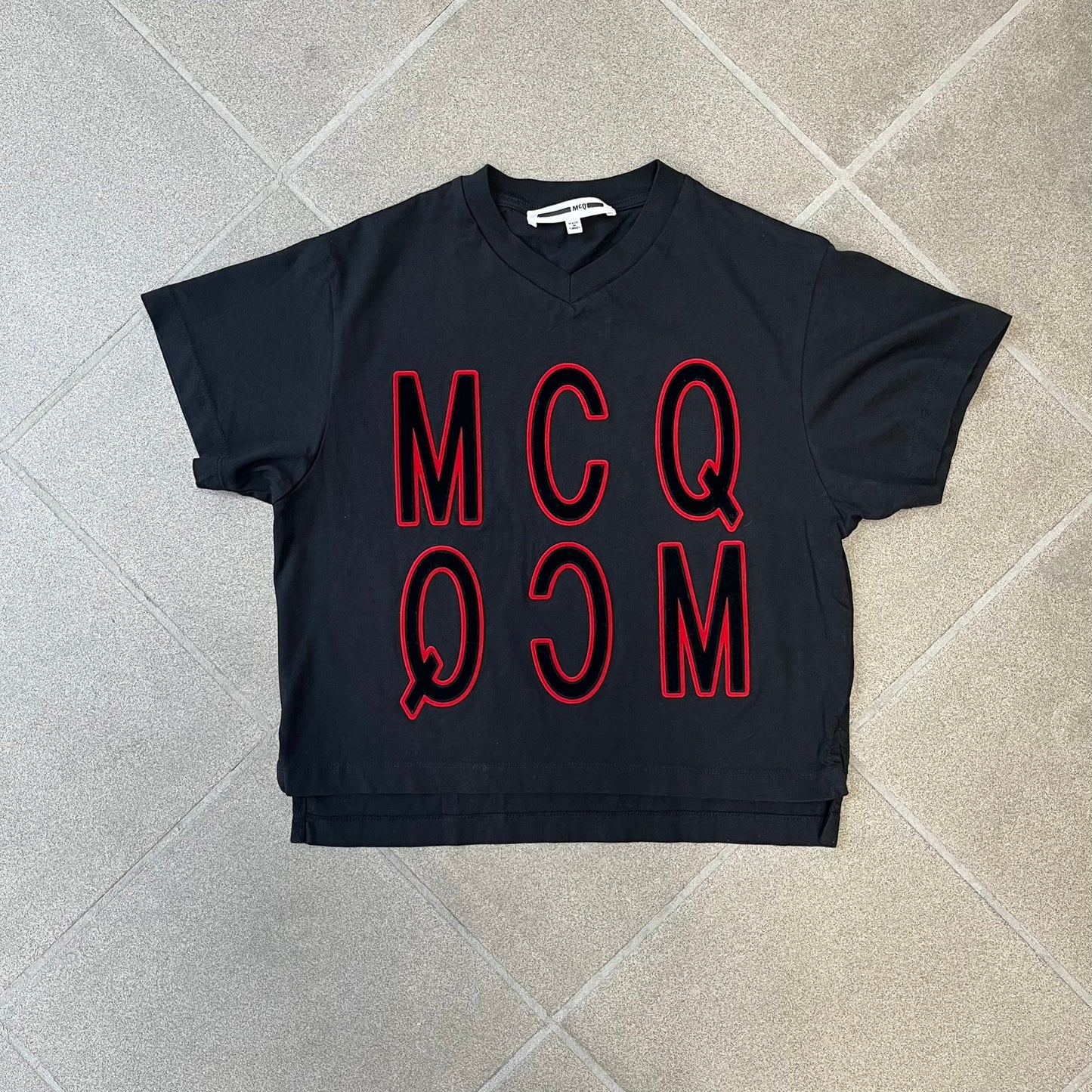 MCQ by Alexander McQueen Velvet Reverse Logo Tee features a black cropped silhouette with a slightly longer back. The front has a large black velvet MCQ logo with a red outline, accompanied by two MCQ text logos: one in the standard orientation and the other reversed as “QCM.”
