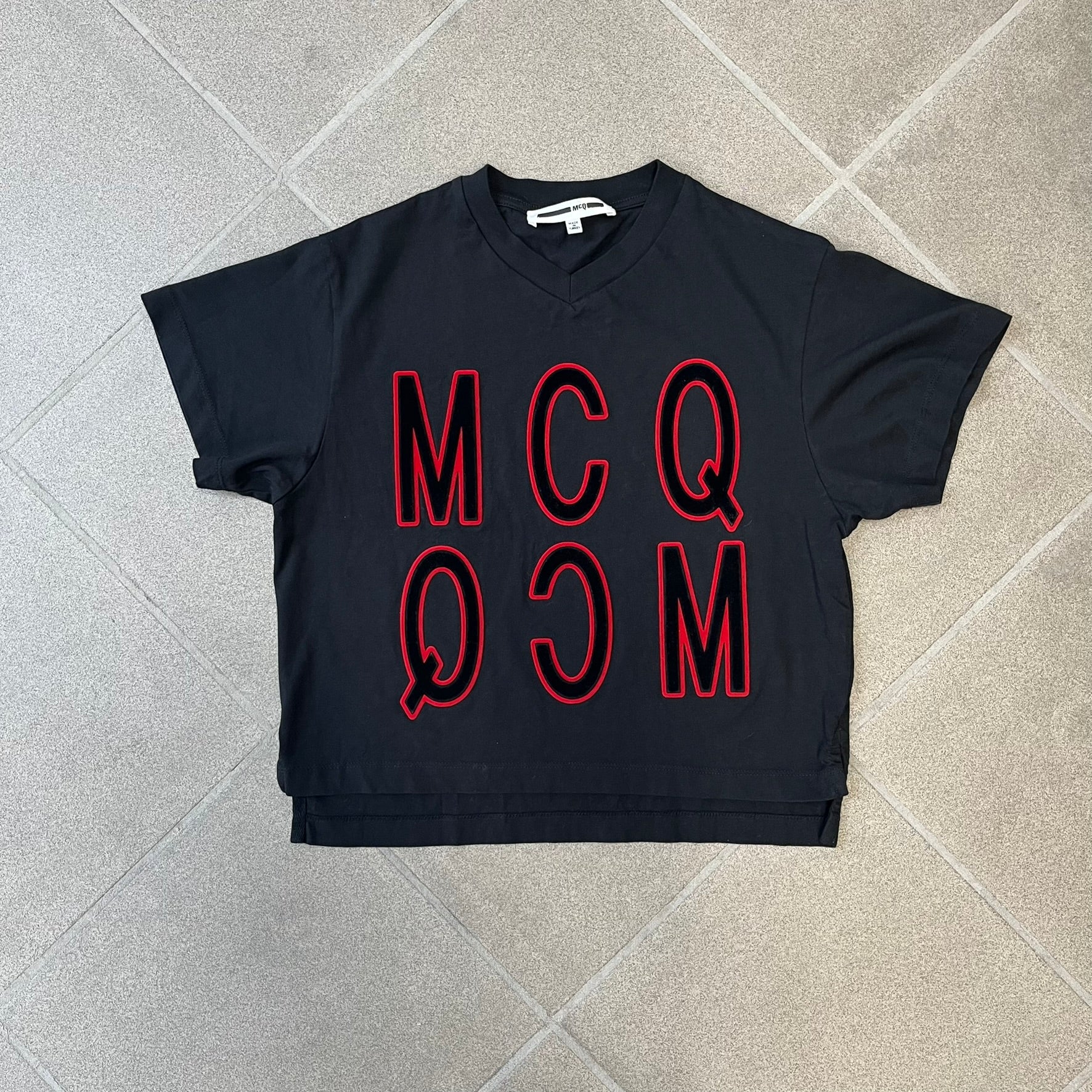 MCQ by Alexander McQueen Velvet Reverse Logo Tee features a black cropped silhouette with a slightly longer back. The front has a large black velvet MCQ logo with a red outline, accompanied by two MCQ text logos: one in the standard orientation and the other reversed as “QCM.”

