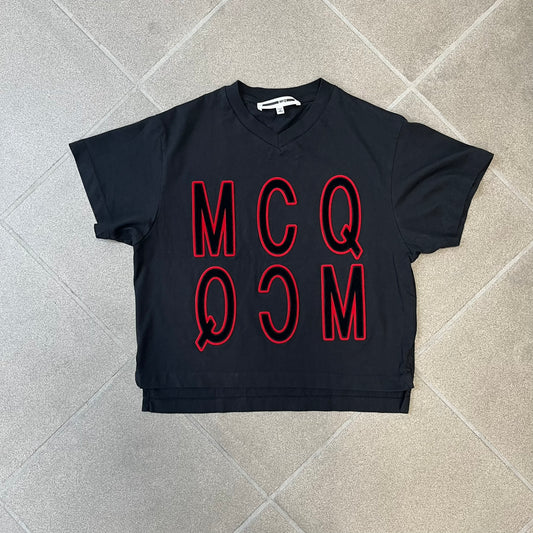 MCQ by Alexander McQueen Velvet Reverse Logo Tee features a black cropped silhouette with a slightly longer back. The front has a large black velvet MCQ logo with a red outline, accompanied by two MCQ text logos: one in the standard orientation and the other reversed as “QCM.”
