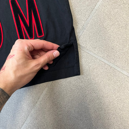 MCQ by Alexander McQueen Velvet Reverse Logo Tee features a black cropped silhouette with a slightly longer back. The front has a large black velvet MCQ logo with a red outline, accompanied by two MCQ text logos: one in the standard orientation and the other reversed as “QCM.”
