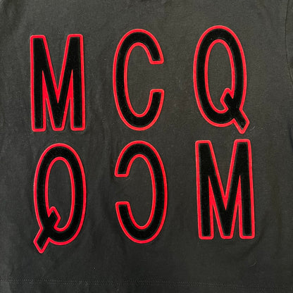 MCQ by Alexander McQueen Velvet Reverse Logo Tee features a black cropped silhouette with a slightly longer back. The front has a large black velvet MCQ logo with a red outline, accompanied by two MCQ text logos: one in the standard orientation and the other reversed as “QCM.”
