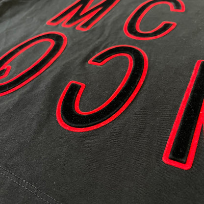 MCQ by Alexander McQueen Velvet Reverse Logo Tee features a black cropped silhouette with a slightly longer back. The front has a large black velvet MCQ logo with a red outline, accompanied by two MCQ text logos: one in the standard orientation and the other reversed as “QCM.”
