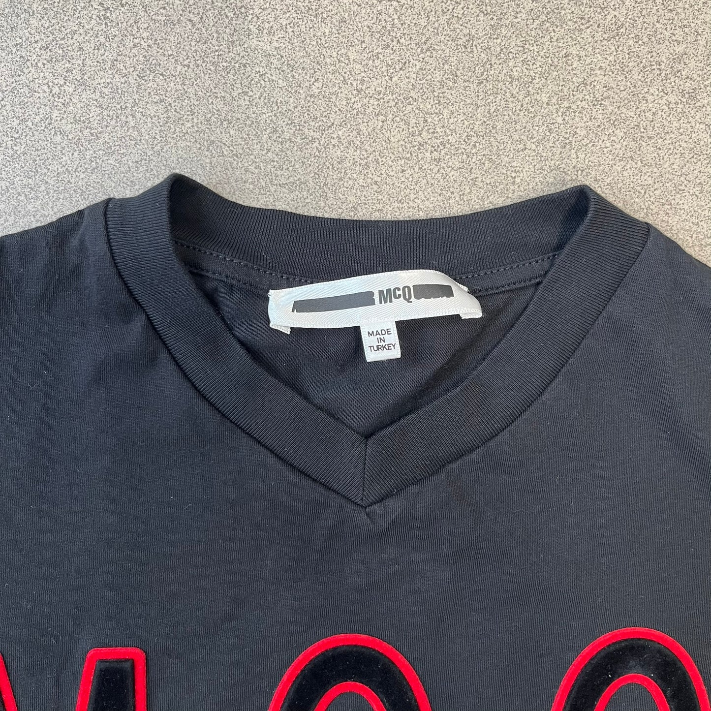 MCQ by Alexander McQueen Velvet Reverse Logo Tee features a black cropped silhouette with a slightly longer back. The front has a large black velvet MCQ logo with a red outline, accompanied by two MCQ text logos: one in the standard orientation and the other reversed as “QCM.”
