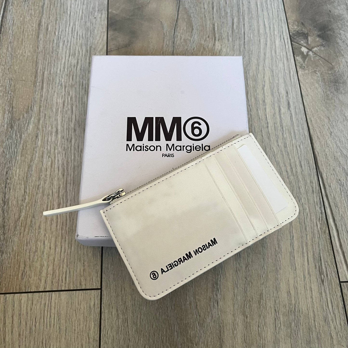 MM6 Maison Margiela White Side Cardholder is a white leather cardholder from MM6 Maison Margiela featuring a horizontal layout with a top zip compartment for coins. The side includes four card slots, while the front displays a mirrored Maison Margiela 6 logo. The item comes with original box and tags.