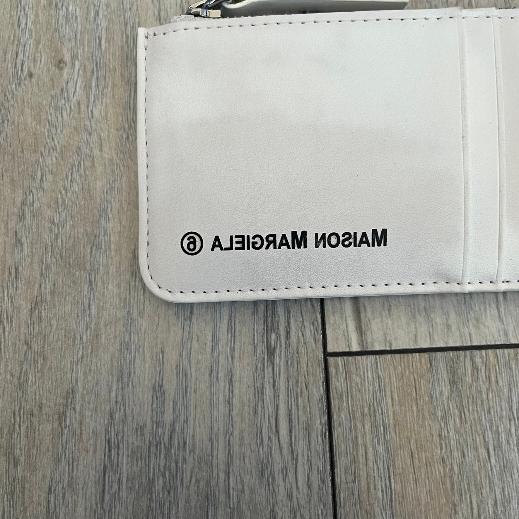 MM6 Maison Margiela White Side Cardholder is a white leather cardholder from MM6 Maison Margiela featuring a horizontal layout with a top zip compartment for coins. The side includes four card slots, while the front displays a mirrored Maison Margiela 6 logo. The item comes with original box and tags.