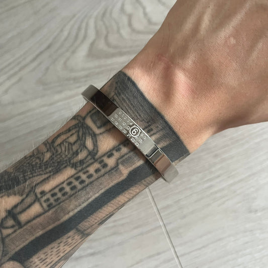 Silver bracelet on a wrist with tattoos, against a light wooden floor background