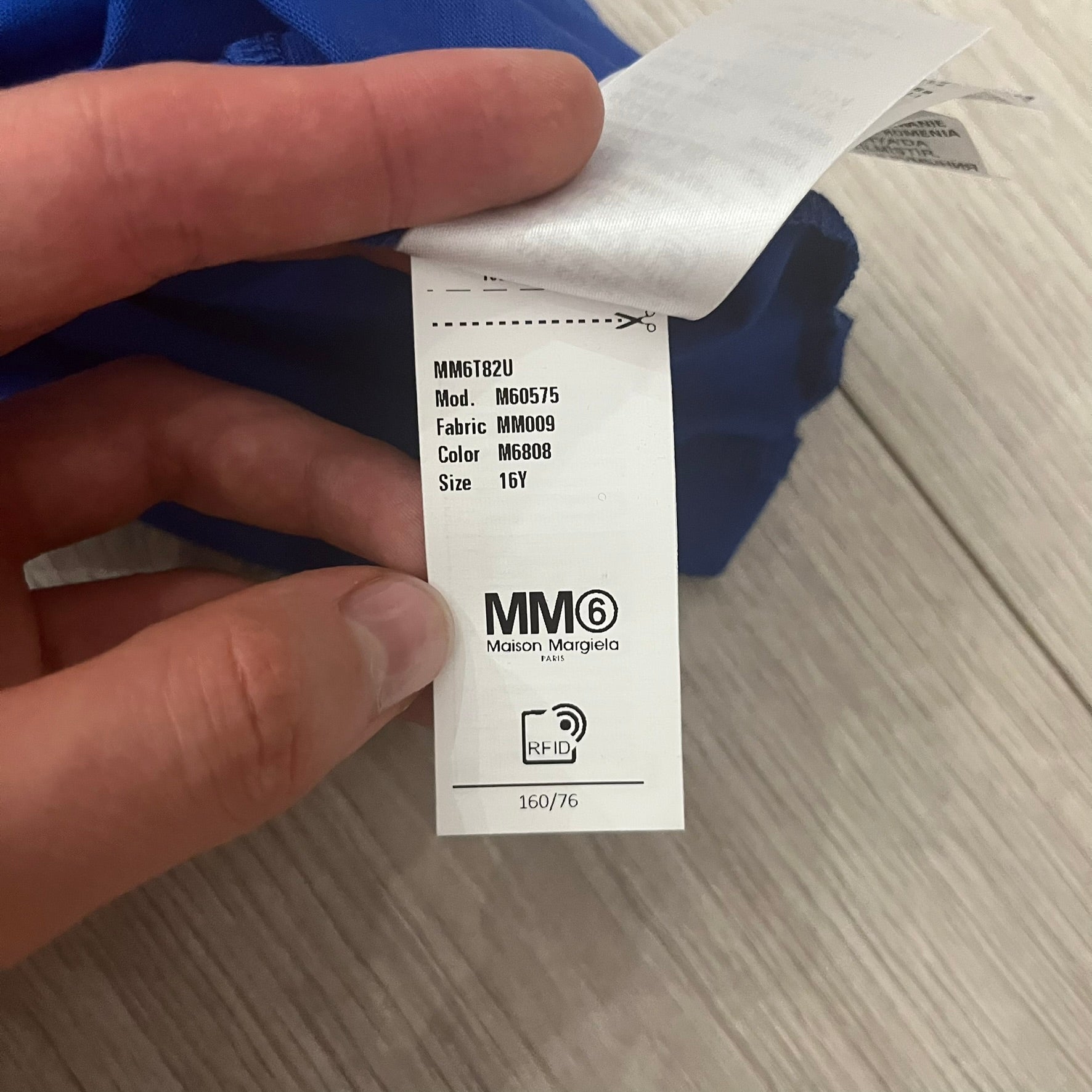 Hand holding a clothing tag with MM6 branding on a wooden surface