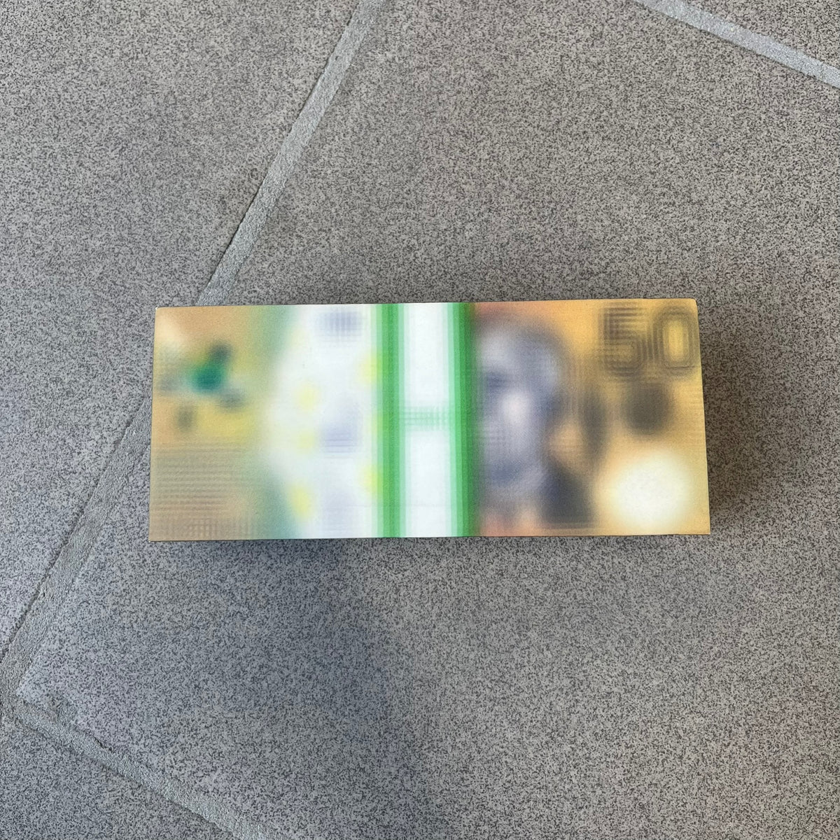 MSCHF Blur A$50 AUD Figure released on May 24rd 2022. The collectible object is designed as a stack of 50 Australian Dollars banknotes with a blurred sculptural effect across the entire surface. The piece recreates the appearance of a money stack while distorting the printed details, giving it a block-like form resembling a stylized or game-like currency bundle.